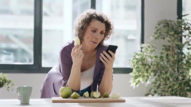 Güzel olgun bir kadının evde akıllı telefon kullanırken elma yediği bir video.
