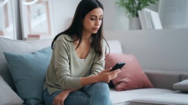 Evdeki kanepede oturan endişeli genç bir kadının cep telefonunu kullanırken çekilmiş videosu.