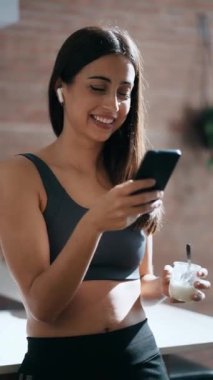 Evde, mutfakta iyogurt yerken cep telefonu kullanan genç bir kadının videosu.