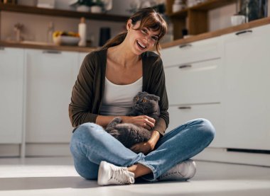 Güzel bir kadının sevimli hayvanlarıyla oynadığı bir fotoğraf. Evdeki mutfakta yerde oturuyor..