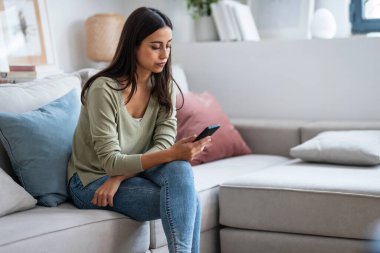 Evdeki oturma odasındaki kanepede oturan sorunlarını düşünürken endişeli genç bir kadının cep telefonunu kullanması..