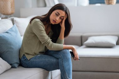 Evdeki oturma odasındaki kanepede oturan sorunlarını düşünürken endişeli genç bir kadının cep telefonunu kullanması..