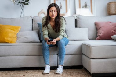 Evdeki oturma odasındaki kanepede oturan sorunlarını düşünürken endişeli genç bir kadının cep telefonunu kullanması..