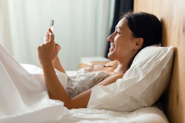 Evdeki yatakta yatarken akıllı telefonuyla video görüşmesi yapan güzel bir genç kadının fotoğrafı.