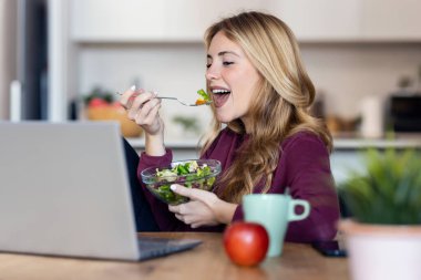 Evdeki mutfakta salata yerken dizüstü bilgisayarla video görüşmesi yapan güzel bir genç kadın..