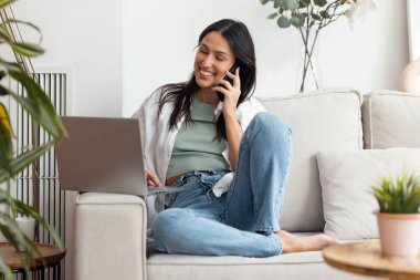 Güzel bir kadının dizüstü bilgisayarıyla evdeki bir kanepede otururken akıllı telefonuyla konuşması.