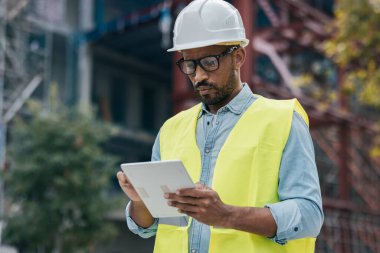 Bir apartman bloğu inşa etmek için gerekli materyalin dijital tabletini taşıyan işçi miğferli bir adamın el yapımı fotoğrafı.