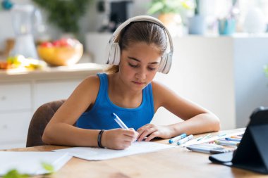 Evdeki mutfakta resim çizerken kulaklıkla müzik dinleyen küçük bir kız resmi.