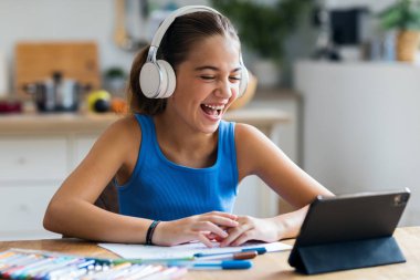 Evdeki mutfakta ödevini yaparken dijital tablet ve kulaklıklarla gülümseyen küçük bir kızın görüntüsü.