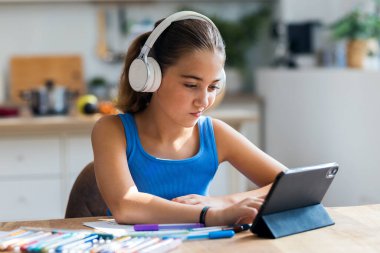 Evdeki mutfakta ödevini yaparken dijital tablet ve kulaklıkla video görüşmesi yapan neşeli küçük bir kız.