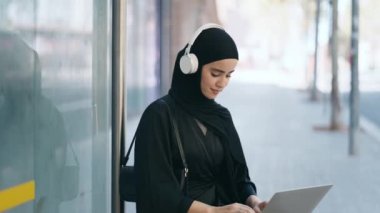 Otobüs durağındaki bankta dizüstü bilgisayarla müzik dinleyen neşeli bir kadının videosu.