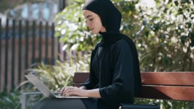 Laptopla çalışan tesettürlü güzel bir kadının şehirdeki bir bankta oturduğu video.