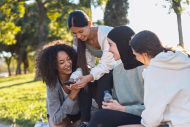 Shot of group of sporty women using smartphone while talking after running while posing at camera in the park clipart