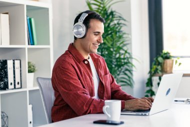 Modern bir girişimde kulaklıkla müzik dinlerken dizüstü bilgisayarla çalışan mutlu girişimcinin fotoğrafı.