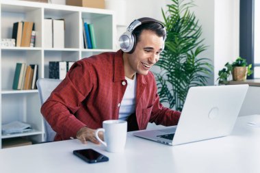Modern bir girişimde kulaklıkla müzik dinlerken dizüstü bilgisayarla çalışan mutlu girişimcinin fotoğrafı.
