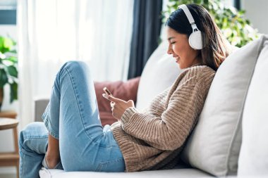 Evdeki kanepede otururken akıllı telefonuyla müzik dinleyen gülümseyen genç bir kadın..