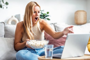 Öfkeyle bağıran ve dizüstü bilgisayarında film seyreden genç bir kadının evdeki kanepede oturup patlamış mısır yerken görüntüsü.