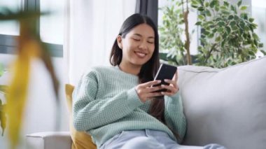 Evdeki kanepede otururken akıllı telefonuyla video görüşmesi yapan güzel bir kadın.