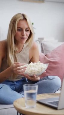 Dizüstü bilgisayarında film izleyen genç bir kadının evde oturmuş patlamış mısır yerken videosu.