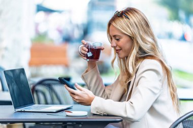 Bir kahve dükkanının terasında akıllı telefon kullanırken dizüstü bilgisayarıyla çalışan çekici genç bir kadının fotoğrafı..