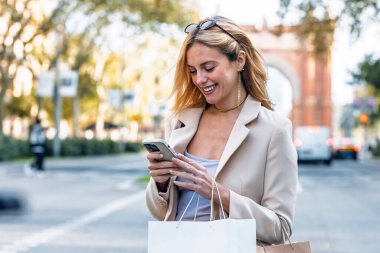 Şehirde alışveriş torbaları taşırken akıllı telefon kullanan mutlu bir kadın.