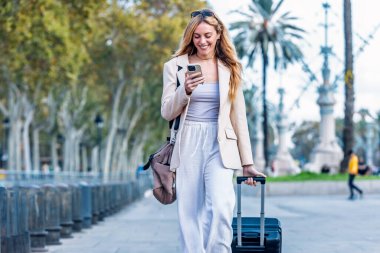 Şehirde akıllı telefonunu kullanırken güzel bir genç kadının bavuluyla yürümesi.