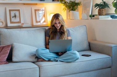 Öğleden sonra evdeki kanepede otururken dizüstü bilgisayarıyla çalışan güzel bir genç kadının fotoğrafı..