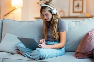 Öğleden sonra kanepede otururken kulaklık takan güzel bir girişimci kadının laptopla çalışırken çekilmiş fotoğrafı.