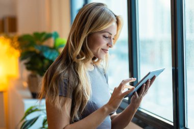 Öğleden sonra pencerenin yanında dikilirken dijital tablet kullanan güzel girişimci kadının fotoğrafı.