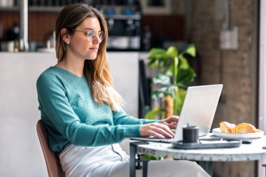 Bir kafede kahvaltı ederken dizüstü bilgisayarla çalışan güzel bir kadının fotoğrafı.