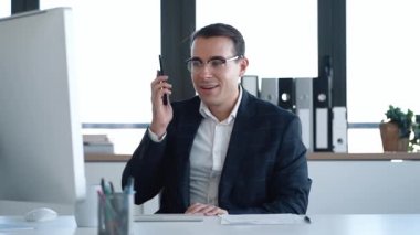Ofiste bilgisayarla çalışırken cep telefonuyla konuşan neşeli iş adamının videosu.