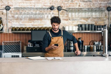 Yakışıklı bir garsonun kahve dükkanında akıllı telefon kullanırken görüntüsü.