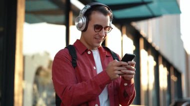 Şehirde kulaklıkla müzik dinlerken akıllı telefon kullanan mutlu bir adamın videosu.