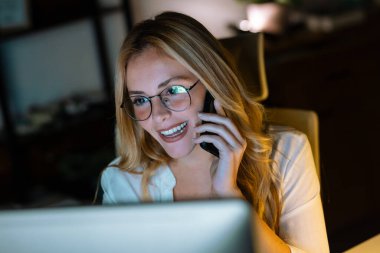 Geceleri ofiste laptopuyla çalışırken gülümseyen bir iş kadınının cep telefonuyla konuşması.