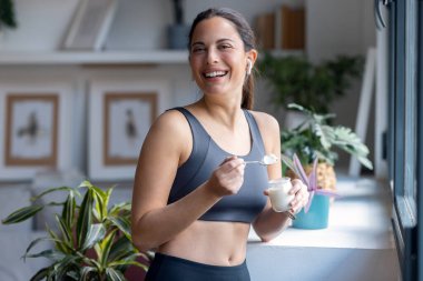 Evde pencerenin yanında duran kulaklıkla müzik dinlerken yoğurt yiyen güzel, sportif bir kadının fotoğrafı..