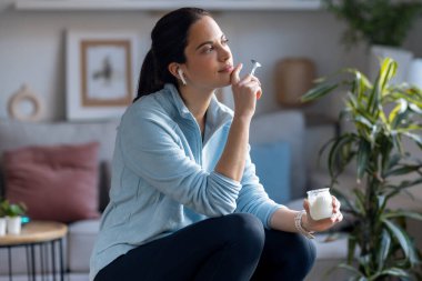 Evde pencerenin yanında oturmuş kulaklıkla müzik dinleyen güzel, sportif bir kadının yoğurt yemesi..