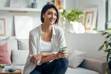 Taburede oturmuş evde kameraya bakarken güzel bir kadının kahve içmesi.