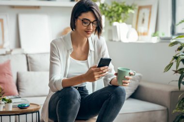 Güzel bir kadının evdeki taburede oturup kahve içerken akıllı telefon kullanması.