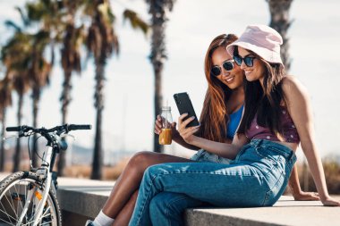 Sahildeki bankta oturup eğlenirken iki mutlu arkadaşın akıllı telefonuyla selfie çekmesi.