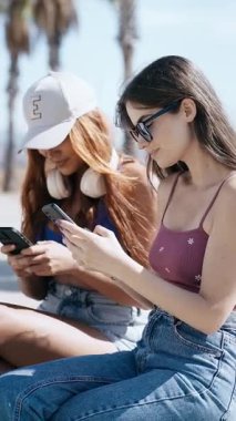 Sahildeki bankta oturmuş meyve suyu içerken akıllı telefonlarını kullanan iki güzel kadının videosu.