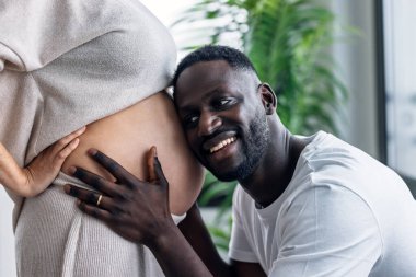 Gülümseyen bir adamın kulağını hamile karısının karnına dayadığı fotoğraf.