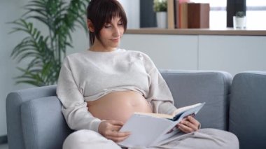 Evdeki kanepede dinlenirken kitap okuyan güzel hamile bir kadının videosu.