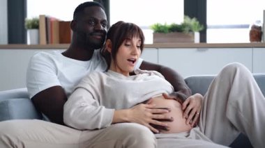 Evdeki kanepede uzanmış güzel hamile bir kadının karnına dokunan yakışıklı adamın videosu.