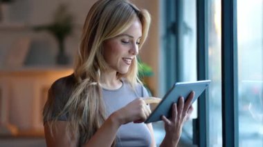 Öğleden sonra pencerenin yanında dikilirken dijital tablet kullanan güzel girişimci kadının videosu.