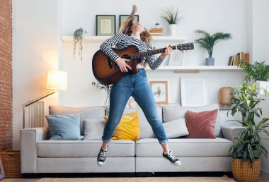 Evdeki oturma odasında dans ederken ve zıplarken güzel bir kadının şarkı söyleyip gitar çalması.