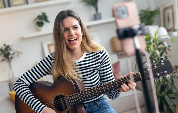 Yemek odasında kendini akıllı telefonuyla kaydederken gitar çalan ve tıngırdatan güzel bir kadın fotoğrafı.