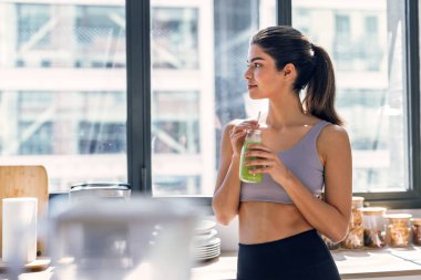 Evdeki mutfakta sebze yeşili el yapımı smoothie içen genç bir kadın..
