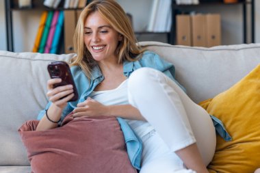 Evdeki koltukta oturan genç ve güzel bir kadının cep telefonunu kullanması.