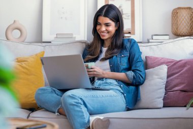 Evdeki kanepede oturmuş bir fincan kahve içerken dizüstü bilgisayarıyla çalışan genç bir kadının fotoğrafı.. 