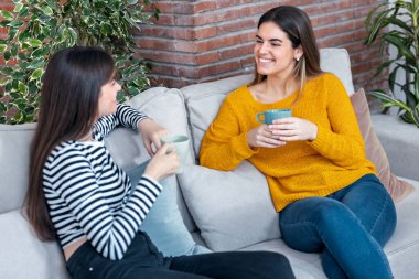 Oturma odasındaki kanepede oturmuş kahve içerken gülümseyen iki genç kadının görüntüsü.
